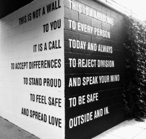 A corner of a black and white wall reads This is not a wall, this is a welcome to you, to every person. It is a call today and always to accept differences, to reject division, to stand proud, and to speak your mind. To feel safe, to be safe, and spread love outside and in. 